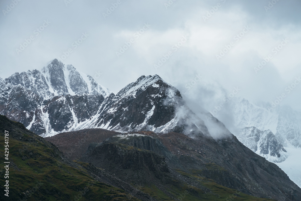 Atmospheric alpine landscape with great mountain peaked top with snow in low clouds. Dramatic mountain scenery with sharp pinnacle in overcast weather. Awesome view to snowy pointy peak in cloudy sky.