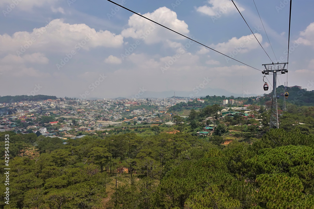 The cable car runs over the coniferous forest. In the distance, in the valley, town houses are visible. There are picturesque cumulus clouds in the blue sky. Dalat. Vietnam