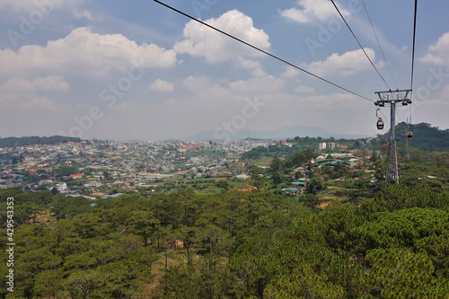 The cable car runs over the coniferous forest. In the distance, in the valley, town houses are visible. There are picturesque cumulus clouds in the blue sky. Dalat. Vietnam