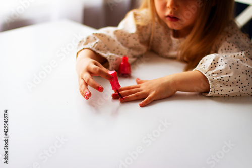 Little girl paints her nails with pink nail polish. Cute toddler girl playing with colorful nail polish doing manicure. Cosmetics and beauty concept.