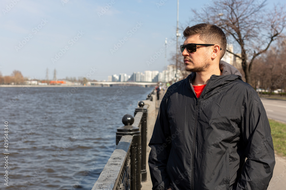 A man in dark sunglasses stands on the banks of a river in the city