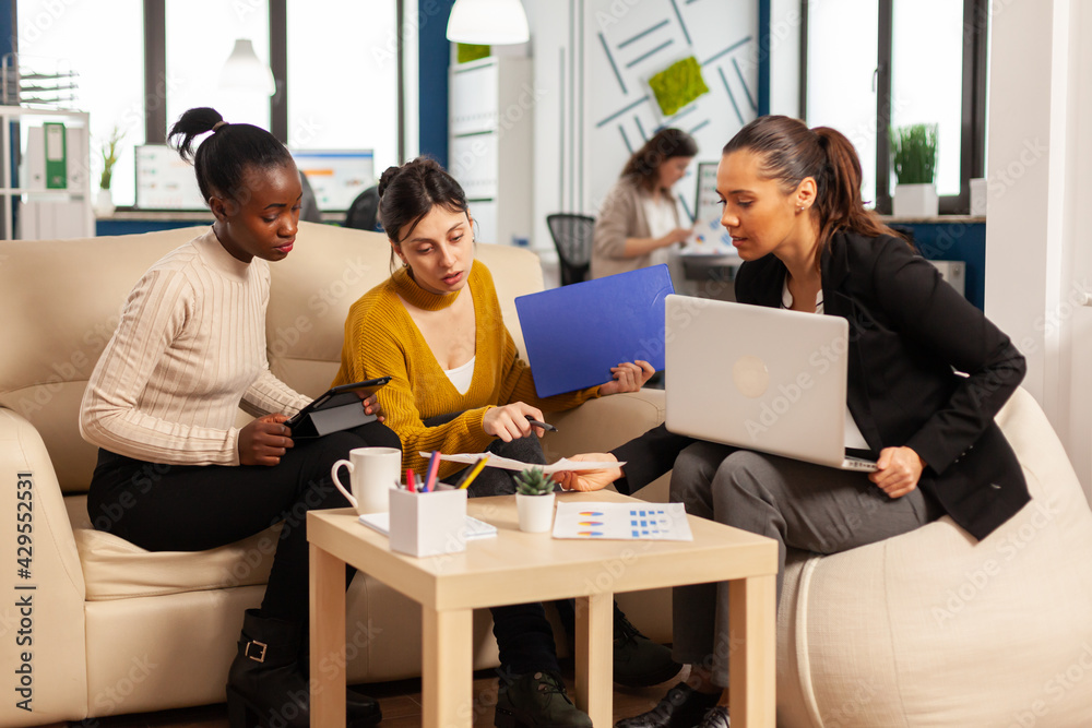 Diverse group of business women sitting on couch in modern corporate startup company office talking about start up financial project and strategy management, using laptop computer and graphics Diverse group of business women sitting on couch in modern corporate startup company office talking about start up financial project and strategy management, using laptop computer and graphics