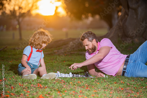 Father and son playing chess lying on grass at lawn park. Fathers Day, love family, parenthood, childhood concept. Active people having fun and playing chess at park, spend time with child.
