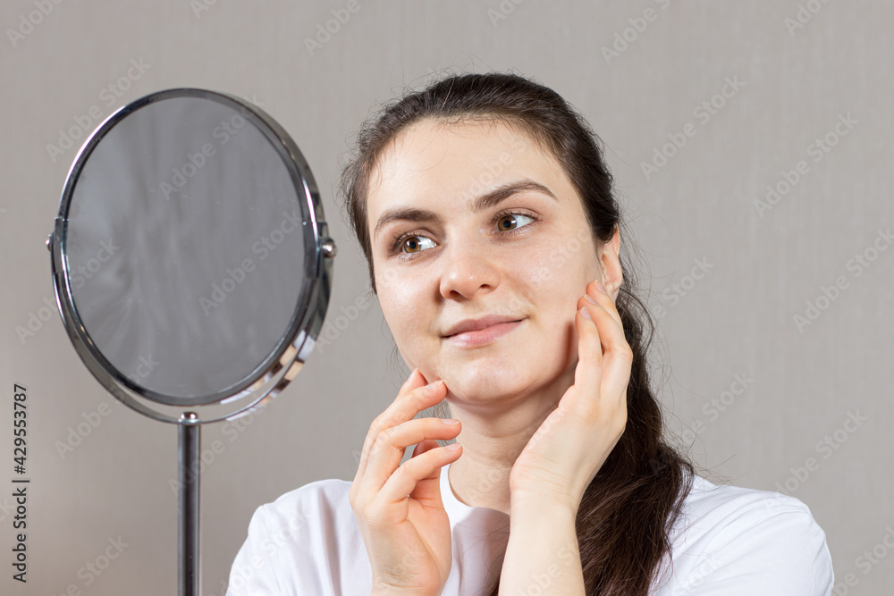 A beautiful young brunette woman 20-30 years old looks in the mirror and admires herself. Self-love, facial skin care. Healthy facial skin for advertising in dermatology and cosmetology A beautiful young brunette woman 20-30 years old looks in the mirror and admires herself. Self-love, facial skin care. Healthy facial skin for advertising in dermatology and cosmetology