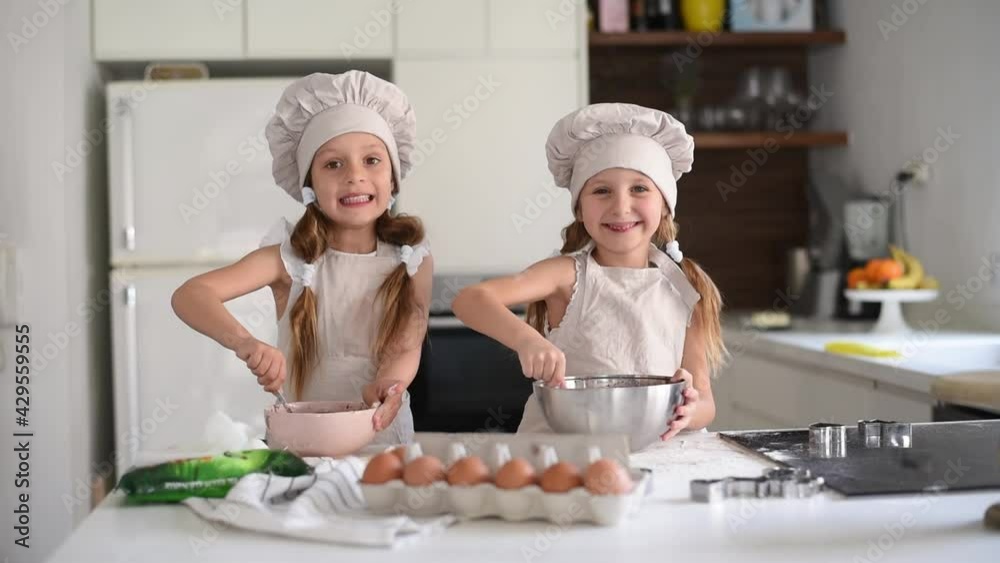 Two small cute girls with amazing smile in special clothes and funny chef cap dance, enjoy, talking, cooking on kitchen cake. Kids make cookie at home, make fun. Children make dessert with eggs Two small cute girls with amazing smile in special clothes and funny chef cap dance, enjoy, talking, cooking on kitchen cake. Kids make cookie at home, make fun. Children make dessert with eggs