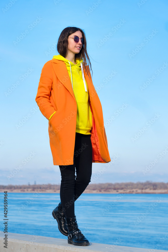 Atmospheric lifestyle photo of young happy woman in sunglasses, orange  coat, yellow hoodie and black jeans walking and relaxing on pier next to  sea. Street Style. Real people concept Stock Photo