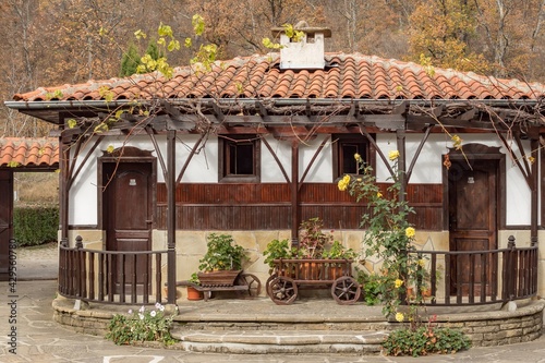 Traditional Bulgarian house exterior, white walls and wooden windows, architecture from Bulgarian National Revival period of the 19th century. Autumn leaves and flowers decoration of yard.
