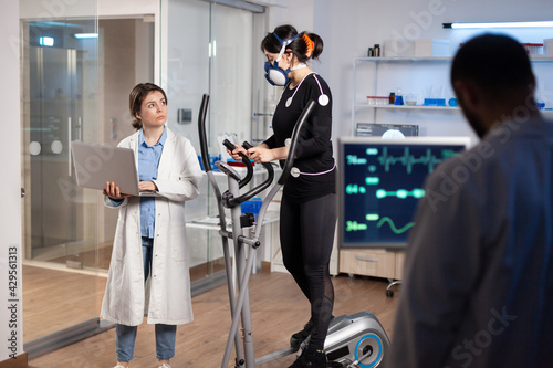 Specialist sport researcher monitoring heart rate on athlete while woman with mask running on cross trainer talking with medical doctor. Physician using laptop controling EKG data in modern laboratory