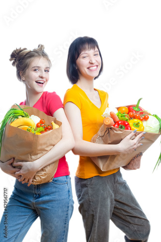 Healthy Lifestyle. Two Young Caucasian Girls Posing With Paper Bags Filled With Grocery And Vegetables Over White Background.