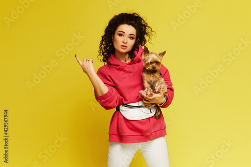 Model girl with afro hair wearing sportswear posing with a small cute dog on her hands over yellow background, isolated