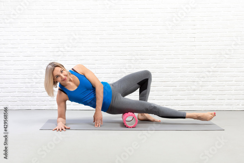 Fit adult caucasian woman in sportswear practice pilates with massage roller under her leg in loft fitness studio indoor.