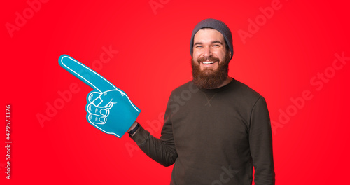 A happy young man is looking at the camera smiling and is holding a big fan glove near a red wall