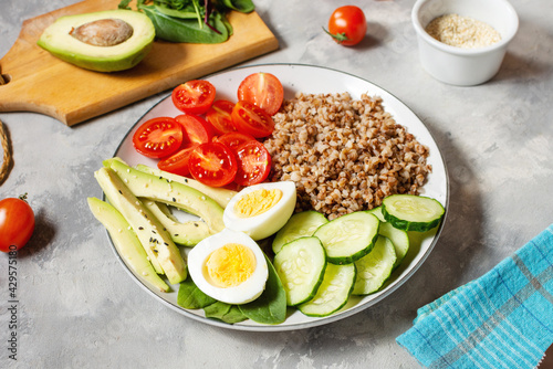 Vegan lunch bowl with avocado, egg, cucumber, tomato and buckwheat on concrete background