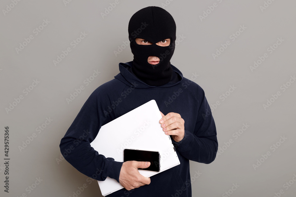 Satisfied man robber steals laptop beech and phone, being dressed in robber mask and black turtleneck, looks at camera firmly holds stolen things, posing isolated over gray background. Satisfied man robber steals laptop beech and phone, being dressed in robber mask and black turtleneck, looks at camera firmly holds stolen things, posing isolated over gray background.