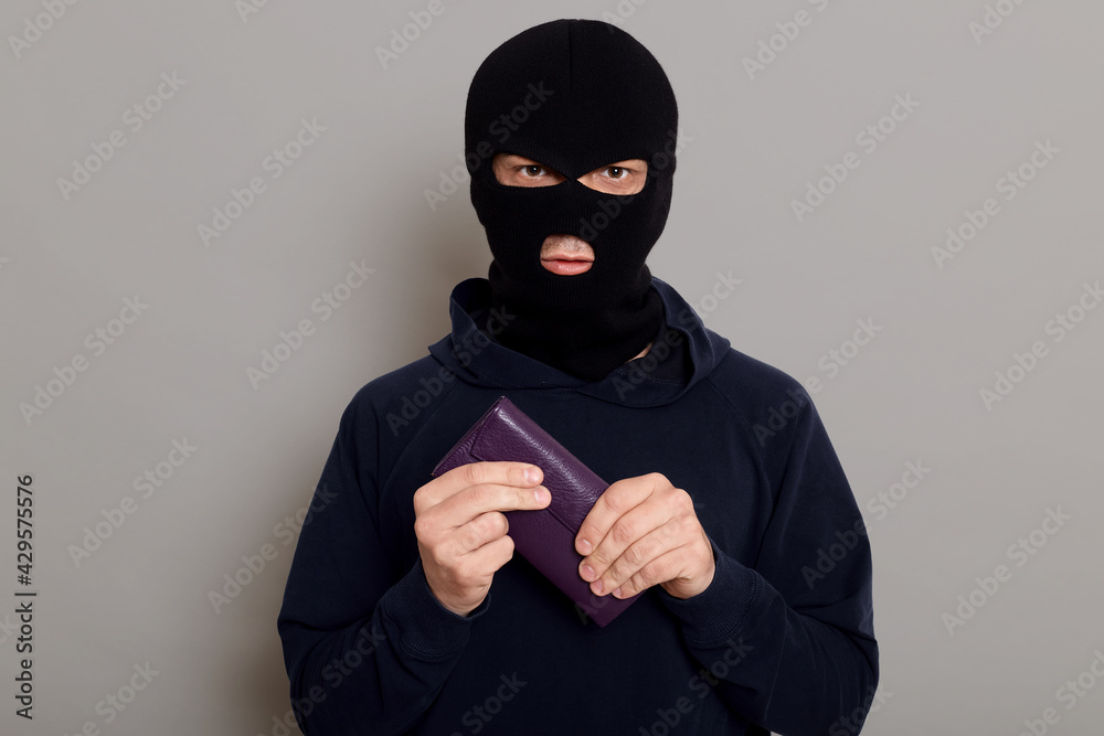 Serious young man burglar in robbery mask and black sweater holds tightly wallet that he just stole, hoping for huge amount of money, standing isolated over gray background. Serious young man burglar in robbery mask and black sweater holds tightly wallet that he just stole, hoping for huge amount of money, standing isolated over gray background.