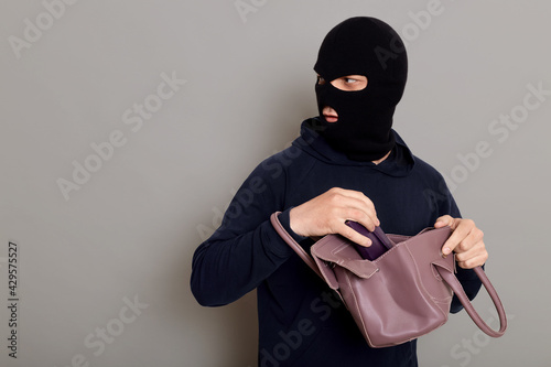 Wallpaper Mural Insidious male villain in balaclava stands with stolen women's bag and wallet, afraid that he will be caught, turns around and looks carefully back, isolated over gray background