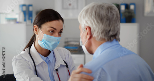 Close up female doctor wearing mask touching senior patient shoulder in clinic office