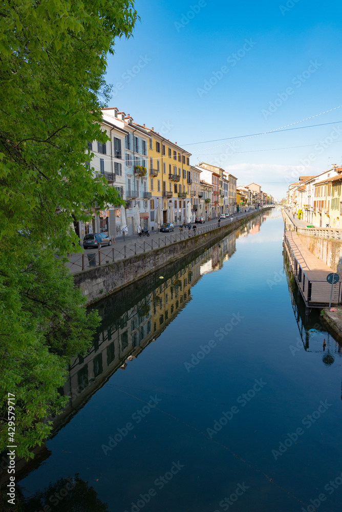 Fototapeta premium Perspective of the famous old Navigli neighborhood in Milan, Italy. Water canal in the foreground, looking like a river, with a green tree. Buildings and blue sky in the background.