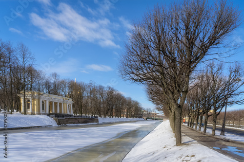 Spring city landscape with a half-frozen river, alleys of age-old branching trees, an old stone pavilion with columns and a blue and white sky. Sunny day in St. Petersburg (Russia). Early spring 