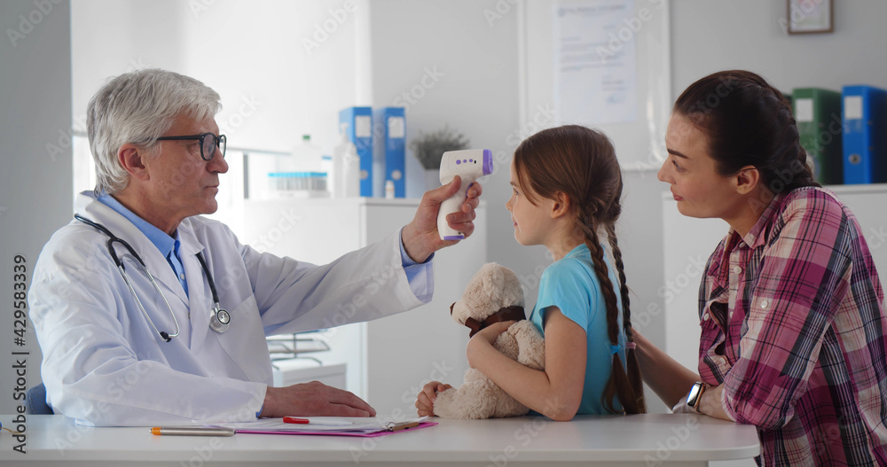 Friendly doctor talking with young woman and daughter measuring temperature with infrared thermometer Friendly doctor talking with young woman and daughter measuring temperature with infrared thermometer
