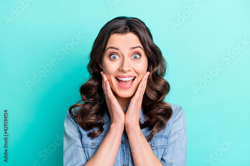 Photo of tricky shocked young woman wear jeans shirt arms cheeks open mouth smiling isolated turquoise color background