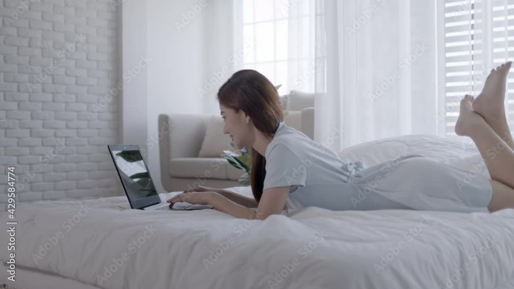 Asian woman working on a laptop sitting on the bed in the house. Asian woman working on a laptop sitting on the bed in the house.