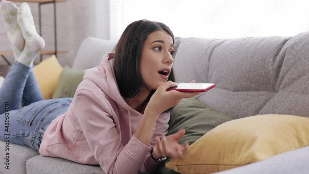 Girl lying on the couch and using voice recognition, dictates thoughts, voice dialing message. Female with smartphone. People lifestyle concept. Woman using smartphone voice recognition in living room
