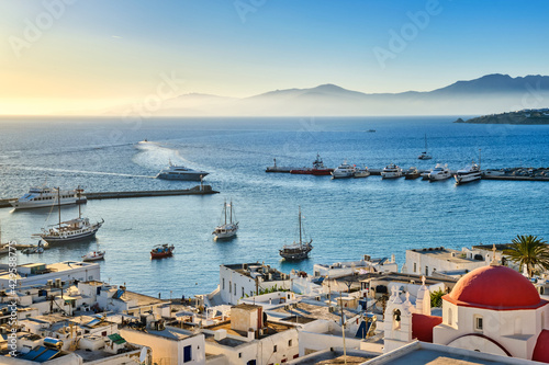 Beautiful view of Chora, Mykonos, Greece at sunset. Port, bay, boats, yachts moored by jetty. Famous whitewashed houses, white church with red dome 