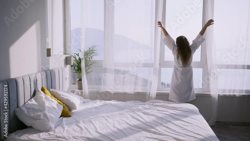 Brunette lady in white bathrobe comes to window to open transparent curtain in spacious hotel room with comfortable bed in sunny morning backside view