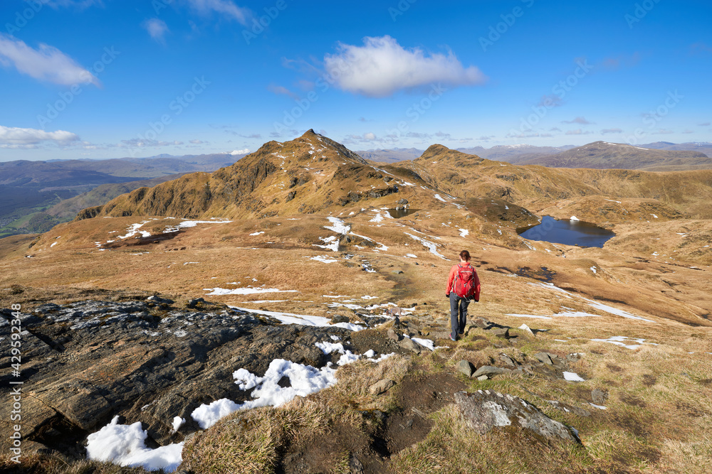 A hiker walking in the mountains towards the summits of Meall Garbh and Beinn nan Eachan from Meall nan Tarmachan in the winter. Scottish Highlands, UK Landscapes.