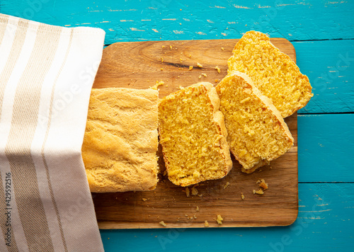 Cornbread is bread containing cornmeal, corn flour. Beautiful homemade yellow sliced cornbread loaf under cotton towel. Blue wood background. Above view. photo