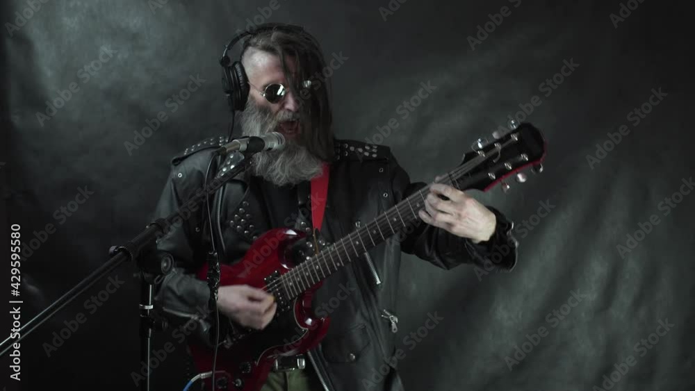 Bearded hairy rocker in headphones with shaved temples, round glasses and leather jacket plays red electric guitar, sings and recording his composition in the studio Bearded hairy rocker in headphones with shaved temples, round glasses and leather jacket plays red electric guitar, sings and recording his composition in the studio