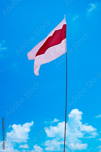  Isolated waving Belarusian national flag on blue sky background with clouds . White-red-white flag. A symbol of peaceful protests in Belarus against the dictatorship after presidential elections.