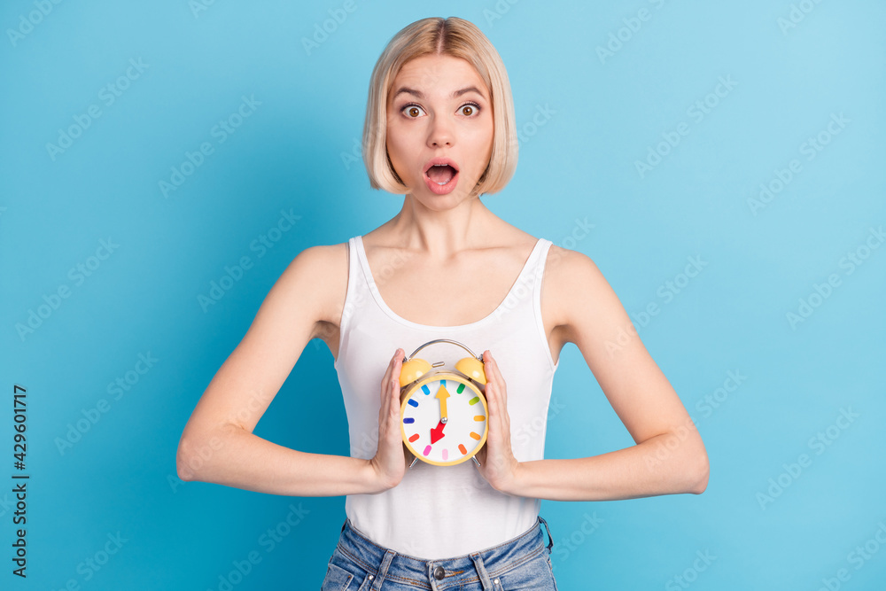 Photo of impressed young girl arms hold classic clock open mouth staring isolated on blue color background Photo of impressed young girl arms hold classic clock open mouth staring isolated on blue color background