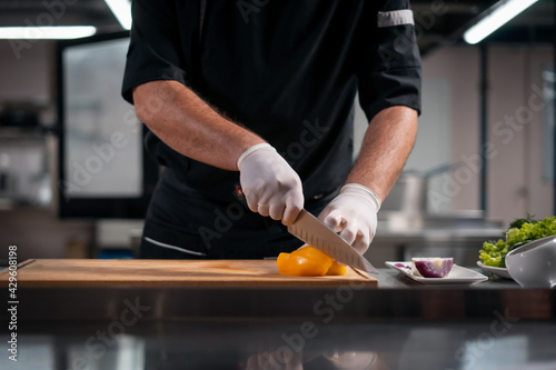close up of chef cook hands in gloves cutting or chop yellow pepper at kitchen