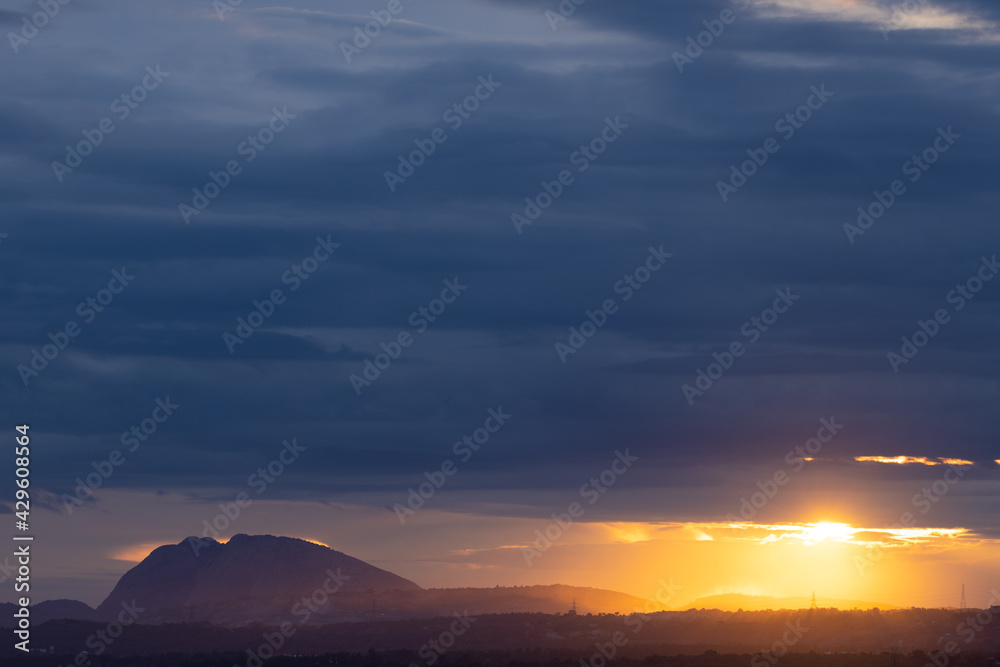 A Panoramic landscape view of hills in the bottom with dark clouds in the horizon and the sun rays passing through them