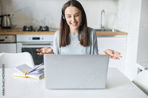 Positive young woman wearing headset talks online, using laptop computer for video connection with colleagues or customers, cheerful female employee works remotely sitting in the kitchen at home