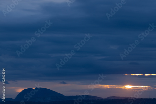 A Panoramic Landscape Silhouette of hills at the bottom with dark clouds in the horizon with sun peeping through them 