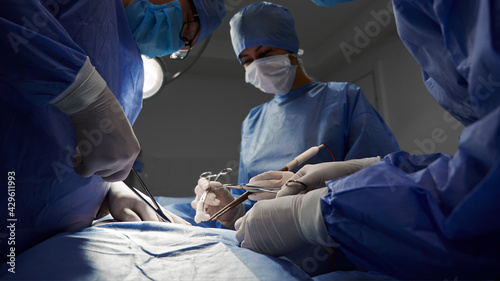 Surgical team in blue medical suits using medical instruments and performing surgical operation. Focus on surgeon hands doing cosmetic surgery at hostpital. Concept of medicine and plastic surgery.