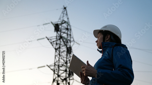 A woman power engineer in white helmet inspects power line using data from electrical sensors on a tablet. High voltage electrical lines at sunset. Distribution and supply of electricity. clean energy