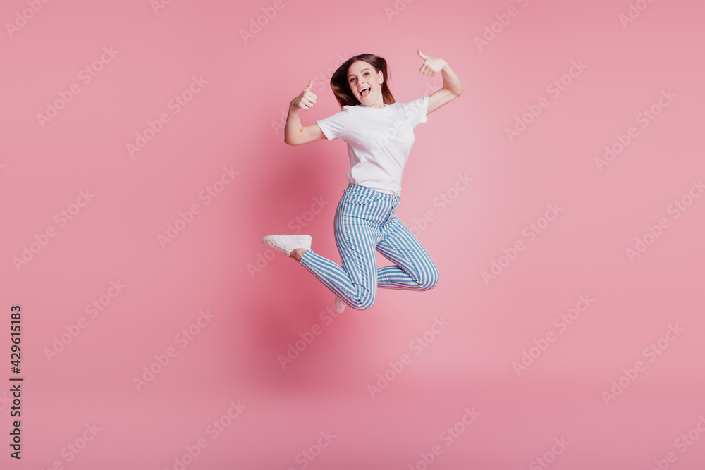 Portrait of playful crazy girl jumping in the air raise two thumbs up on pink background