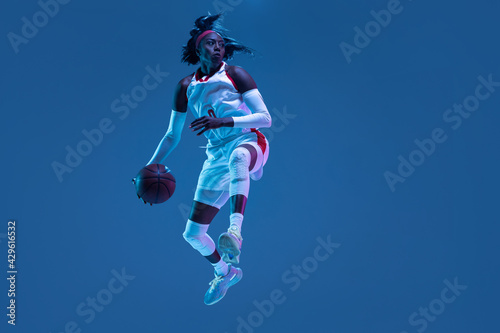 Beautiful african-american female basketball player in motion and action in neon light on blue background. Concept of healthy lifestyle, professional sport, hobby. © master1305