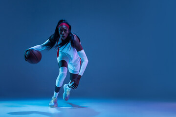 Beautiful african-american female basketball player in motion and action in neon light on blue background. Concept of healthy lifestyle, professional sport, hobby.