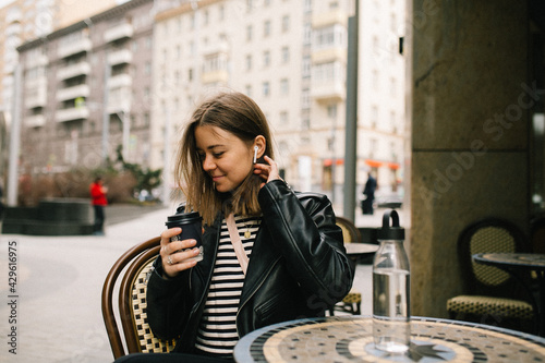 Young pretty girl listening to music in the big city.She is seating at the table. girl in a leather jacket with headphones drinking coffee near business center . smiling and loathing. 