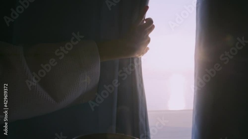 Curly haired lady in white bathrobe opens curtains to look outside large window drinking hot coffee in dark hotel room at sunrise time closeup