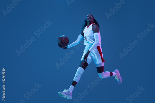 Beautiful african-american female basketball player in motion and action in neon light on blue background. Concept of healthy lifestyle, professional sport, hobby.