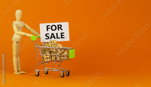 House for sale symbol. Miniature shopping cart with wooden houses, words for sale. Wooden model of human. Beautiful orange background, copy space. Business and house for sale concept.