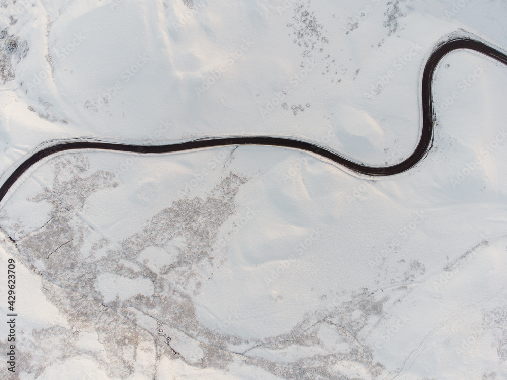 Aerial view of long winding black road through mountain range snow covered landscape. Looking down on single red car driving in icy conditions all alone winter alpine conditions