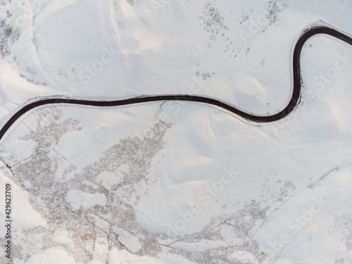 Aerial view of long winding black road through mountain range snow covered landscape. Looking down on single red car driving in icy conditions all alone winter alpine conditions © Matthew
