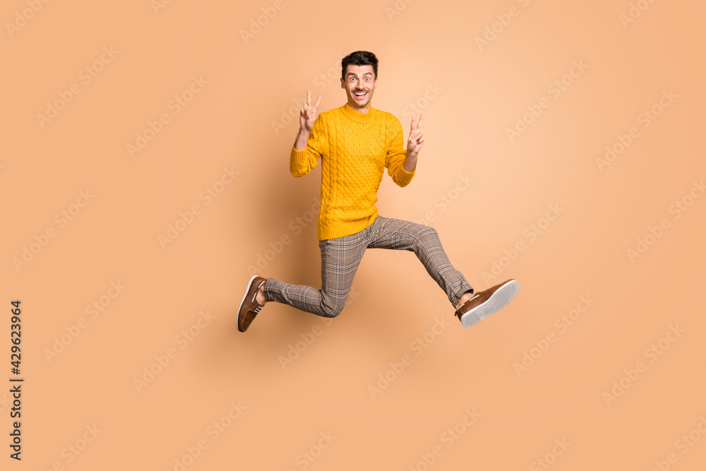 Full length photo portrait of funny man making two v-signs jumping up isolated on pastel beige colored background Full length photo portrait of funny man making two v-signs jumping up isolated on pastel beige colored background